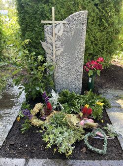 Stehender Stein hergestellt aus hellem Granit mit Stahlkreuz umrandet von Blumen  | © Messerschmidt Gmbh